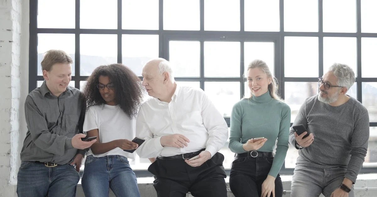 Um grupo multicultural de colegas sentado em ambientes internos, compartilhando smartphones e criando uma atmosfera de equipe amigável. Contexto: