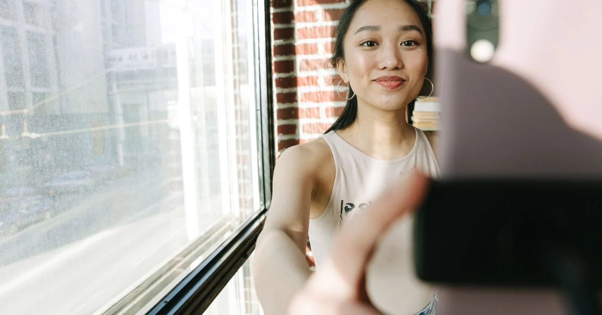 Mulher asiática sorrindo tirando uma selfie perto de uma grande janela com fundo de parede de tijolos. Contexto: Como escolher influenciadores para uma campanha