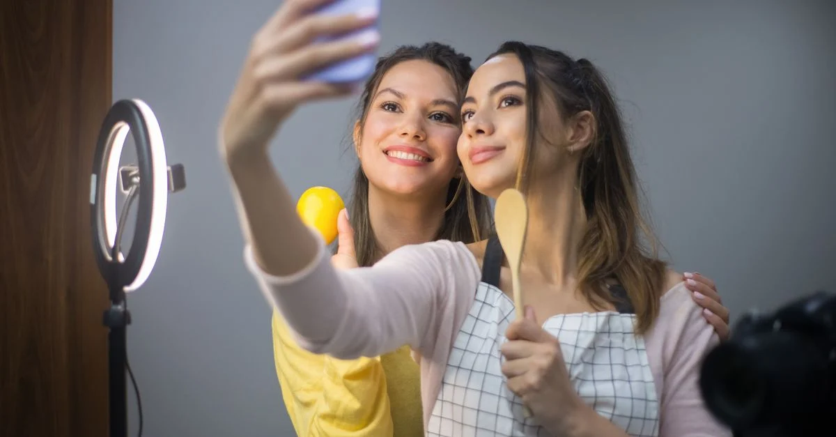 Duas mulheres sorrindo enquanto tiram uma selfie com um anel de luz e smartphone em ambiente interno. Contexto: Direitos de uso em UGC: imagem, mídia paga, prazo e licenciamento