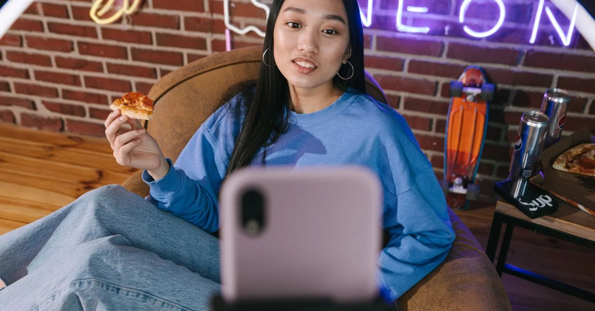 Jovem mulher asiática transmitindo vídeo enquanto saboreia pizza em um ambiente aconchegante com luzes de néon. Contexto: Live commerce: como fazer sua primeira live de vendas
