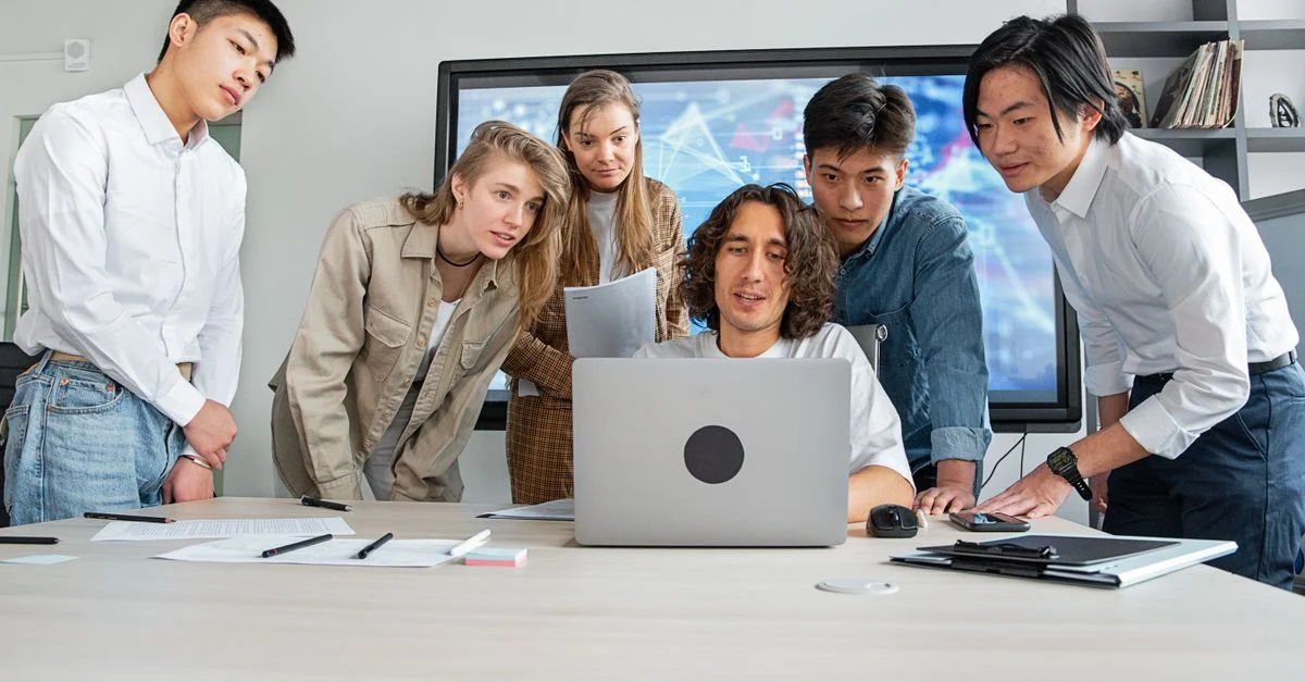 Um grupo diverso de jovens profissionais fazendo um brainstorming durante uma reunião colaborativa em torno de um laptop em um escritório. Contexto: Meta Ads: como anunciar no Facebook e Instagram