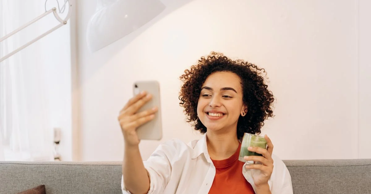 Mulher sentada dentro de casa, tirando uma selfie enquanto segura um smoothie verde, promovendo um estilo de vida saudável. Contexto: Partnership Ads: o que são e como anunciar com creators