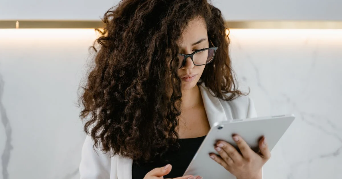 Jovem mulher com cabelo cacheado e óculos usando um tablet dentro de casa para trabalho remoto. Contexto: Quanto custa tráfego pago? Orçamento, gestão e exemplos