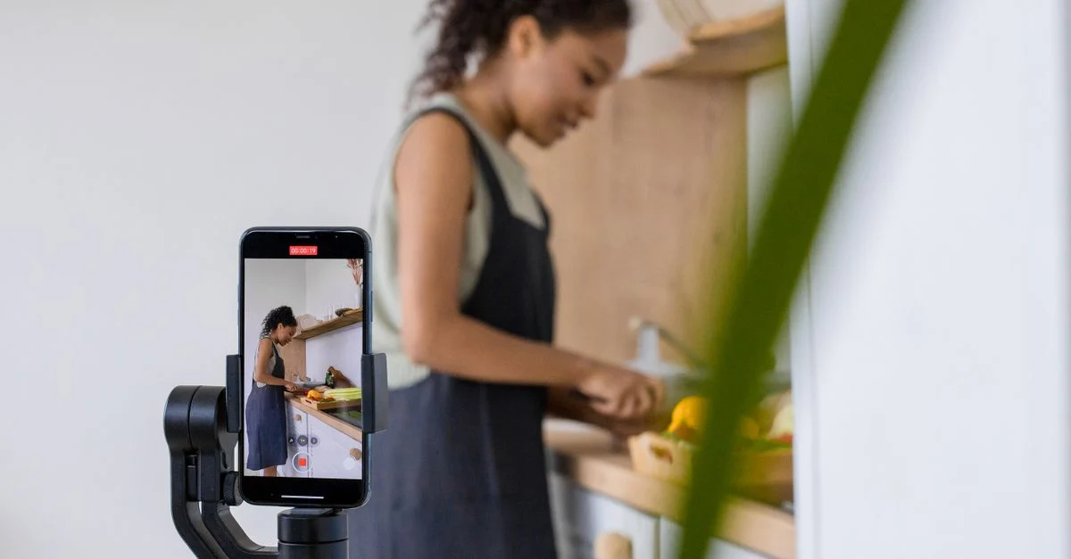 Uma mulher grava um tutorial de culinária em um smartphone com estabilizador. Ambiente de cozinha moderna. Contexto: TikTok Ads: como funciona, formatos e custos