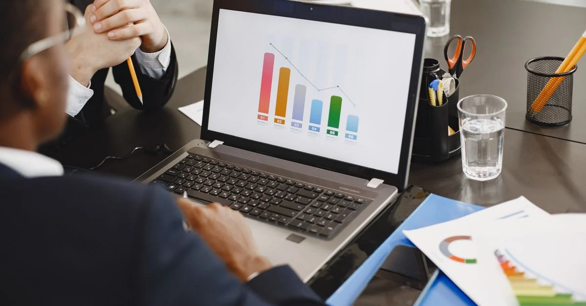 Dois profissionais analisando um gráfico de negócios em um laptop durante uma reunião. Contexto: Tráfego Pago no Google Ads: como funciona e quanto custa