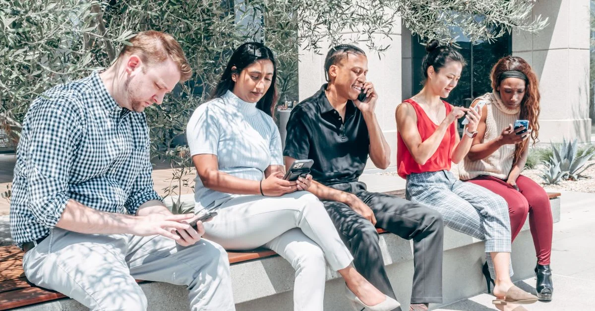 Um grupo diversificado de adultos usando smartphones enquanto estão sentados em um banco ao ar livre. Contexto: Tráfego Pago no Instagram: como funciona e quanto custa